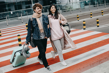 Young modern couple walking on crosswalk. Guy hold hand on suitcase. Attractive brunette walk besides man. Going to airport. Travel abroad.
