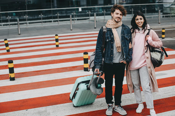 Young positive cheerful couple going to travel. Posing on camera standing on crosswalk. They have suitcase and packpack. Warm clothes. Stand at airport or railway station.