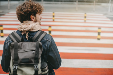 Obraz premium Young attractive man with curly hair stand in front of crosswalk and look to right. Carrying backpack. Walking to airport or railway station. Going abroad or on vacation.