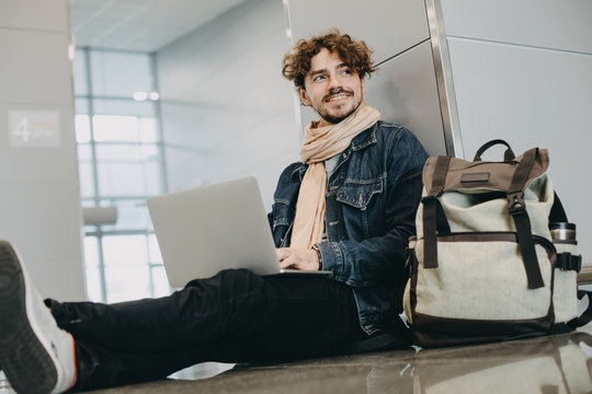Young man with curly hair and backpack sit on floor in airport. Look to side and smile. Hold white laptop on nap. Work remote. Vacation mood or unofficial business trip. Daylight.