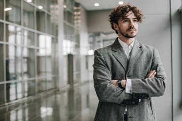 Fototapeta premium Picture of young serious businessman stand alone in room and pose. Hands crossed. Calm peaceful concentrated guy. Expert or diplomat. Empty room. Business day. Wear suit or official jacket.
