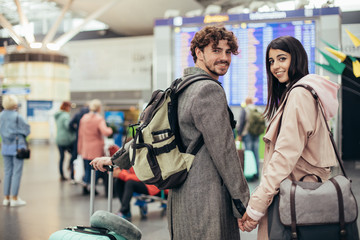 Beautiful young man and woman hold each others hands and look back on camera. Smile. Ready to travel or have trip. Vacation mood. Carry backpack, purse or suitcase. Excited and happy.