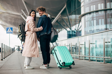 Young couple stand outside of airport and posing. Look back on camera and smile. Carry backpack, purse and suitcase. Woman hold his hand. After vacation of trip. Travel together.