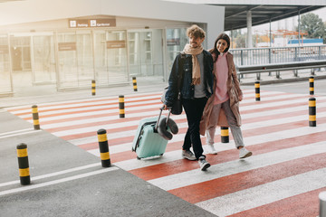 Picture of young man and woman walk on crosswalk together. Guy carry suitcase. Goint to travel abroad. Vacation or trip. Sunlight on picture. In rush to airport.