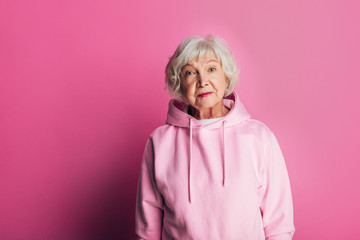Nice quiet old woman posing on camera. Serious and concentrated without smile. Stand alone and wear pink hoody. Beautiful senior woman isolated over pink background.