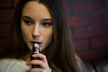 Vaping teenager. Young pretty white caucasian teenage girl with problem skin smoking an electronic cigarette indoors. Life-threatening bad habit. Vape activity.