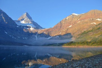 Cold sunny morning in Assiniboine Provincial Park. Lake in front with rolling fog, Mount Assiniboine covered with snow in towering over whole scenery.