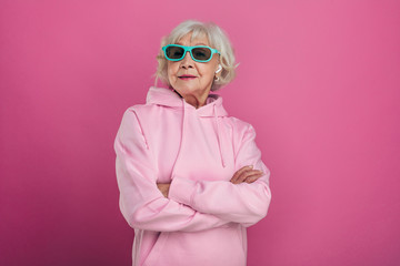 Cool old woman in funny green sunglasses posing on camera. Hands crossed. Wear pink hoody. Stylish modern grandma. Isolated over pink background. - obrazy, fototapety, plakaty