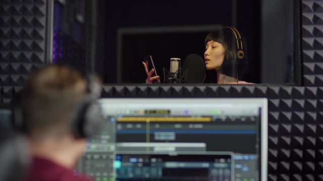 Zoom In Of Young Female Asian Pop Singer In Earphones Looking At Smartphone Screen And Singing In Microphone Standing In Booth While Sound Engineer Recording Her Song In Music Production Studio