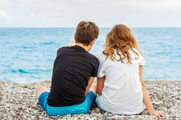 Couple of beautiful teens, first love. Guy hugs a girl sitting on the pebble beach next to each other and looking at the sea. Shoot from behind