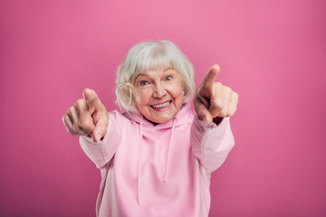 Happy cheerful old woman point forward on camera and smile. Wear stylish modern sweater. Stand alone and pose on camera. Cool grandma. Isolated over pink background.