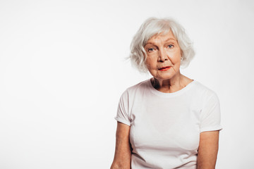 Old woman with white hair and wrinkles on face. Sit alone and pose on camera.Look straight without smile. Wear white shirt. Isolated over white background.