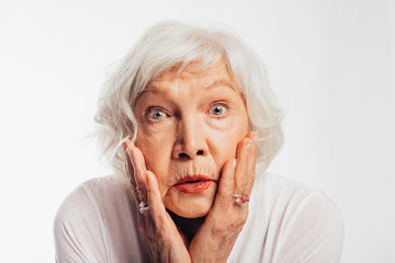 Amazed emotional old woman look straight on camera with wonder. Hold hands on cheeks. Playing perfomance alone. Female elder with grey hair and red lips isolated over white background.