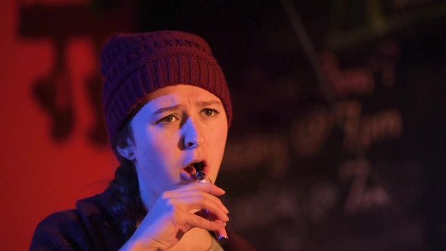 Young women speaks seriously on stage and smokes a vape pen before walking off to the right