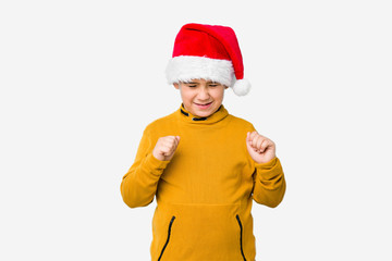 Little boy celebrating christmas day wearing a santa hat isolated raising fist, feeling happy and successful. Victory concept.