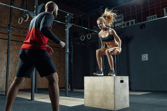 Young fitness woman is doing exercises with the help of a personal trainer in a modern gym Trainer helping woman on her work out routines. Sporty girl doing a box jump at the gym A couple of athletes.
