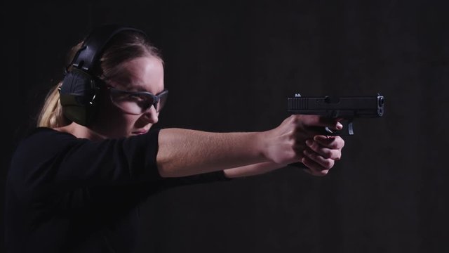 Slow motion shot of pretty blonde caucasian woman with ear and eye protection, aiming and firing a blowback pistol with recoil, in calm, focused manor, side view with dark background.