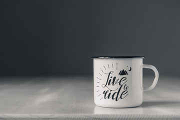 Enamel white metal mug with black line on the edge with the inscription on white wooden table on gray background. Place for text or advertising