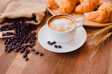 Top view white hot coffee cup and beans on wooden table with croissant bread background.Latte art coffee menu for breakfast in the coffee shop.Selective focus.