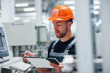 Making notes. Industrial worker indoors in factory. Young technician with orange hard hat