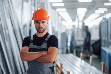 Stands with arms crossed. Portrait of male industrial worker indoors in factory. Young technician...
