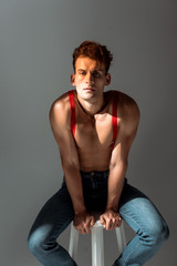 shirtless man in red suspenders looking at camera and sitting on white chair isolated on grey