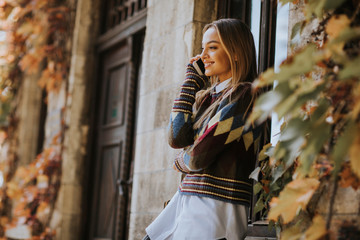 Fototapeta premium Pretty young woman standing with mobile phone on street at autumn day