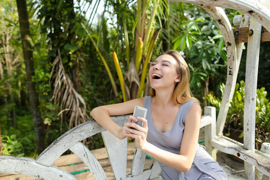 Young smiling woman riding on swing and using smartphone in exotic garden, palms in background.