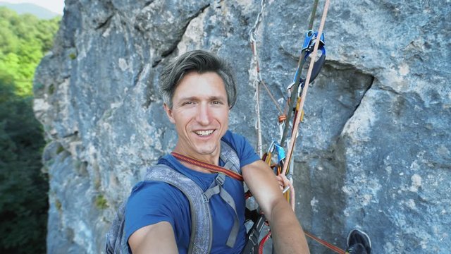 smiling man photographer capturing video selfie while hanging on climbing ropes on the cliff. man hanging using climbing gear, ascender and belaying devices. sport climbing shooting in nature