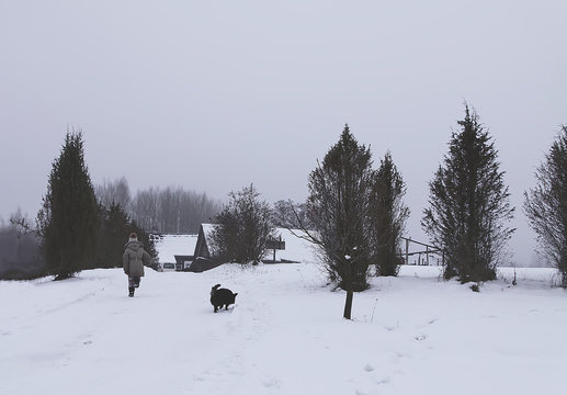 Little girl child in a beige coat running with small balck dog on snow covered field in the village