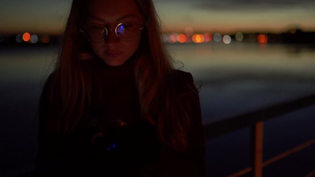 Sad young woman using her phone texting and standing on a pier - Vivid night on a river with bokeh light in the background - 1.4 aperture thin dop
