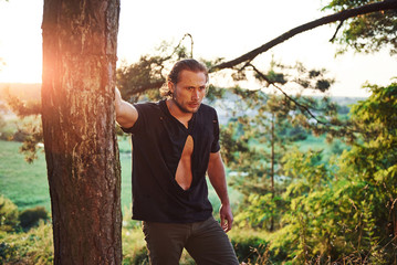 Leaning on the tree. Amazing sunlight. Handsome man with muscular body type is in the forest at daytime