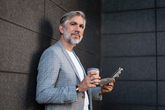 Fashionable mature, businessman leaning at a wall with newspaper and takeaway coffee