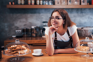 Leaning on the wooden table. Young female cafe worker indoors. Conception of business and service