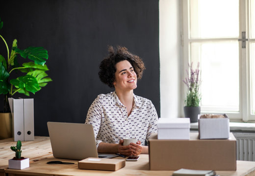 Smiling young woman sitting at desk with coffee cup, laptop and packages
