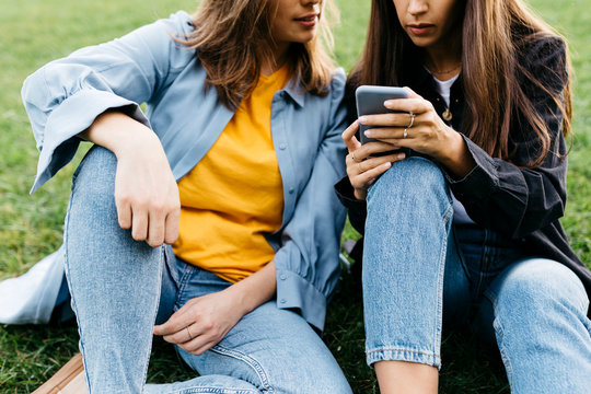Two friends looking on the smartphone, sitting on a meadow of a park