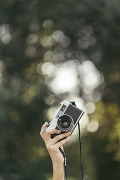 Hand Holding Grey And Black Analog Camera With Forest Bokeh Effect In Background