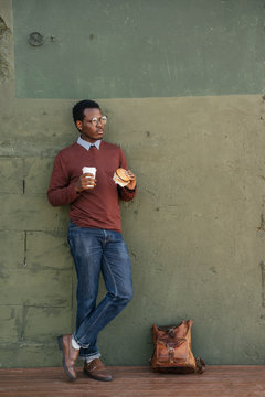 Young man with a cheeseburger and coffee to go, leaning on wall