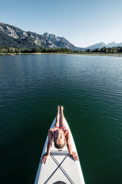 Girl sitting on SUP board and looking at camera, from above, Lake Bannwaldsee, Germany