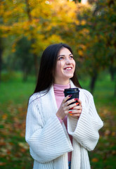 young female in white cardigan in the autumn park with black cup of coffee