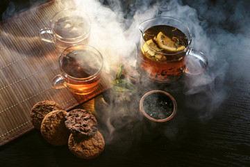Cafe or tearoom hookah lounge concept. Two cups of aroma herbal black or red tea with some fresh cookies with spices closeup on wooden table in clouds of smoke.