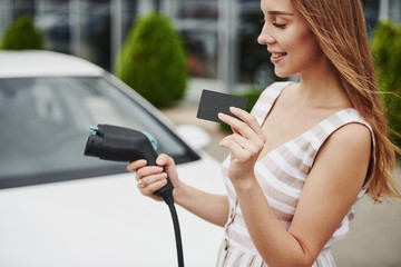 Holds black card. Woman on the electric cars charge station at daytime. Brand new vehicle