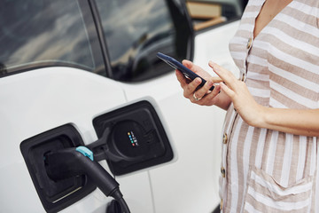 Using smartphone while waiting. Woman on the electric cars charge station at daytime. Brand new vehicle