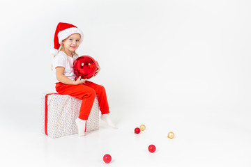 Little smiling girl in santa helper hat sitting on a giftbox with red ribbon and holding red christmas ball, white isolated background. Christmas, winter, happiness concept.