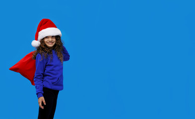 Fototapeta na wymiar Little girl with cute smling wearing santa hat carrying gift bag isolated on blue background. Winter holidays, Christmas. Copy space. Horizontal view.