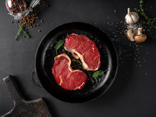 Raw steaks with seasonings in iron pan, black background. Fresh meat, herbs, garlic, champignon, pepper, salt, wooden board. Close-up. Top view.