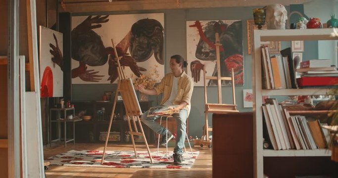 Caucasian good looking young man artist painting a picture while sitting at the easel in the comfortable nice art studio.