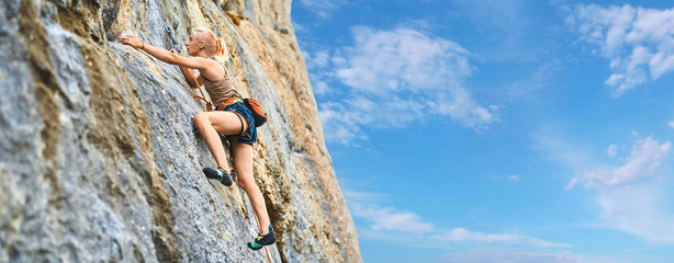 side view of young athletic woman rock climber climbing on the cliff. a woman climbs on a vertical rock wall on the blue sky background