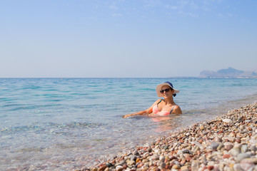 Sexy girl wearing bikini and hat and enjoying water at pebble beach.beautiful woman relaxing at empty pebble beach in natural reserve.well-shaped body in bikini sitting in water, admiring fascinating