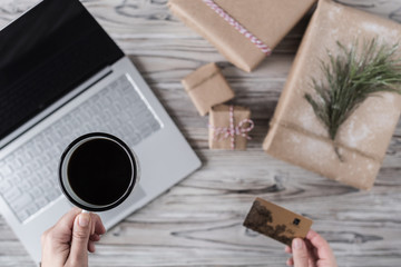 Woman with cup of coffee, credit card and tablet. Packing gifts. Cardboard box in craft paper on the rustic wood planks background. Christmas and New Year concept.